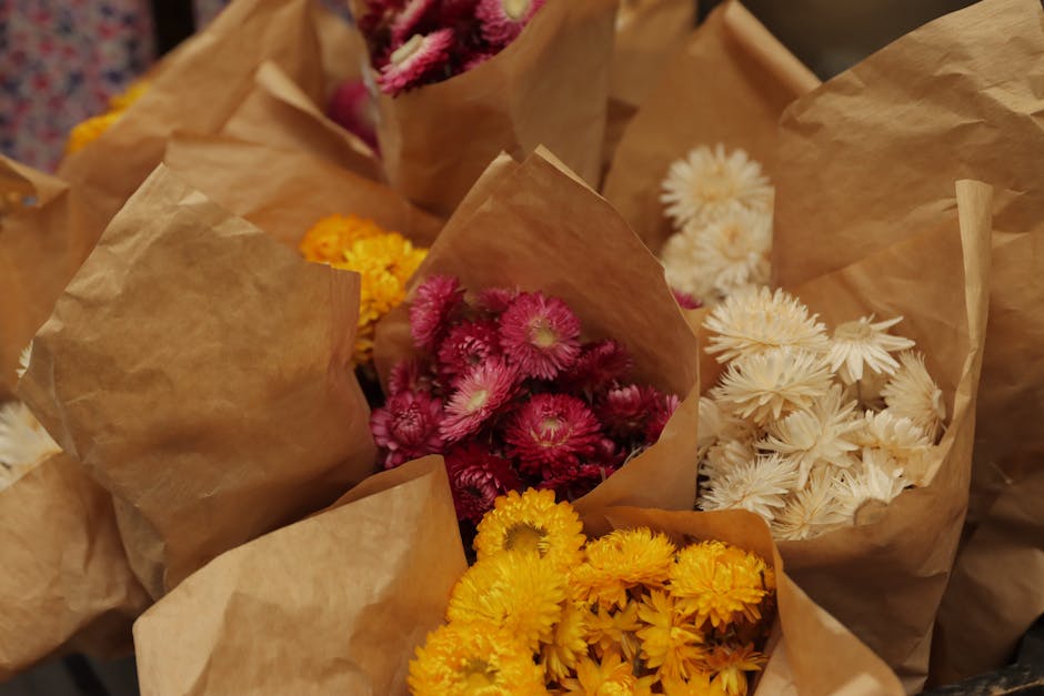 Multiple bouquets of fresh flowers wrapped in brown kraft paper, arranged on a surface. The arrangements include vibrant pink chrysanthemums, bright yellow blooms, and soft white flowers, all with vis