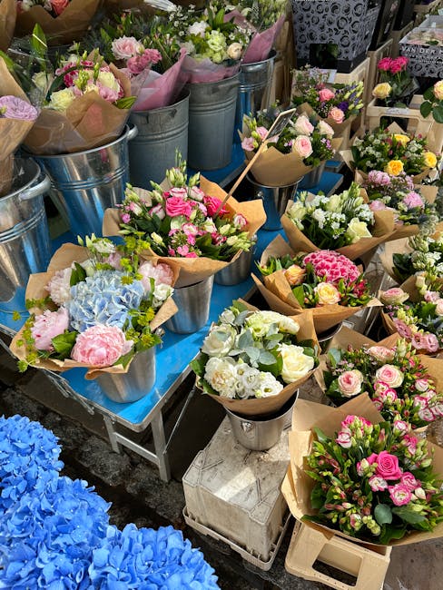 A display of freshly cut flower bouquets arranged in brown kraft paper and placed in metal buckets and vases on a table outside a flower shop. The bouquets feature pastel pink roses, white and pale pi