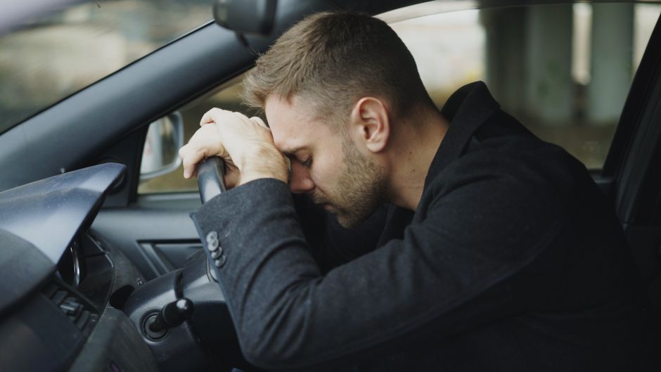A man with short light brown hair and a beard, dressed in a dark suit, sits in the driver's seat of a car, resting his forehead against the steering wheel with a distressed expression. The car's inter