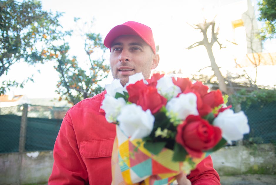 A man dressed in a red jacket and matching red cap holds a bouquet of fresh flowers outdoors. The bouquet features a mixture of red and white blooms, including roses and other blossoms, wrapped in col