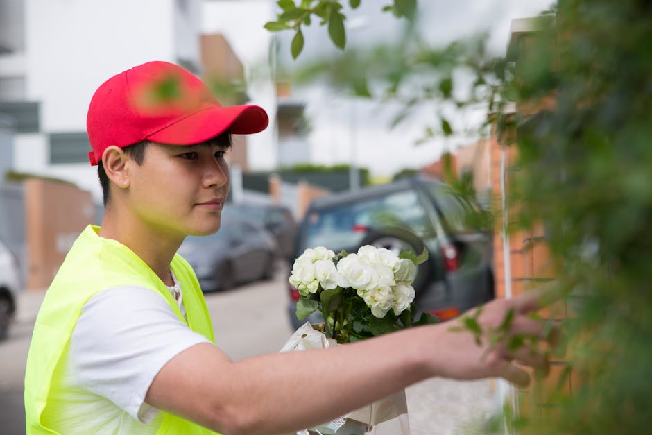 A male florist from flowerspeckham.co.uk wearing a red cap and a fluorescent yellow safety vest is arranging a floral gift of white hydrangeas and roses, carefully holding the bouquet with his left ha