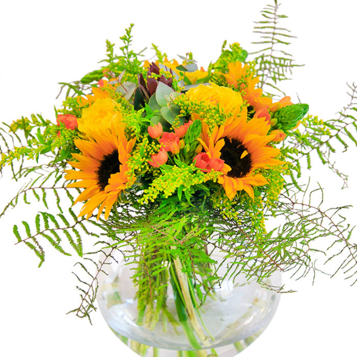 From the first glance, this sun-drenched arrangement radiates pure cheer, alive with the warm spectrum of yellows and oranges so familiar to Peckham in late summer. Towering sunflowers, their velvety golden petals unfurling around espresso-hued centers, form the exuberant core of the bouquet, evoking the full bloom of wild fields you might find beyond Peckham Rye. Interwoven with these are buttery yellow garden roses-each bloom displaying a tightly layered, spiraling elegance-placed with the skilled hand of a true florist. Punctuating the yellows, clusters of glossy, scarlet Hypericum berries lend vivid highlights, their sheen offering playful contrast amidst a backdrop of lush, textural greenery. Cascading ferns bend gracefully at the edges, while touches of soft eucalyptus and light green goldenrod add subtle depth and movement. Every detail is visible through the bowl-shaped glass vase, where the crisp stems are artfully arranged in clear water-a testament to freshness and craftsmanship. In soft midday light, the petals seem to glow, filling the space with a sense of optimism and vivacity. This arrangement is perfect as a celebratory centerpiece for gatherings in Peckham homes, a thoughtful gift for anniversaries, birthdays, or to share a bit of sunlight on a rainy day, hand-gathered with pride by local florists who know the neighbourhood's unique flair.